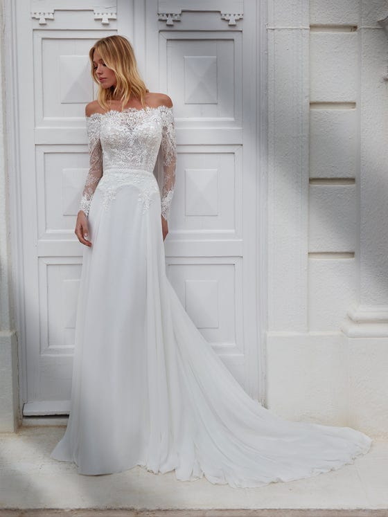 Sensual dress with lace bodice and bateau neckline with exposed shoulders and long semi-transparent sleeves. A design with a flowy chiffon skirt decorated with a guipure waistband. 
