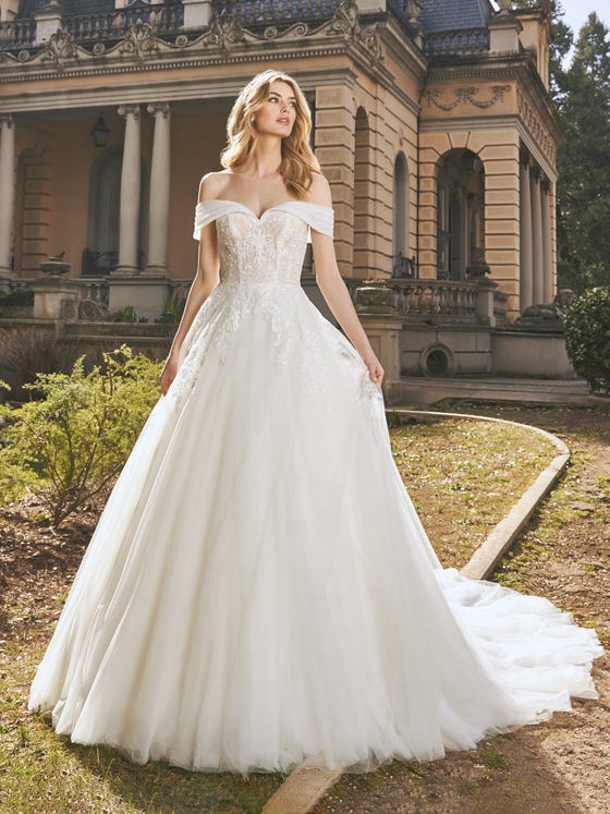 Classicism and romanticism meet in this sensational tulle and lace dress full of sparkle and glimmers. A design with a fitted bodice and a beautiful sweetheart neckline set off by the draped off-the-shoulder sleeves. The decorations cascade down over the top of the skirt. 