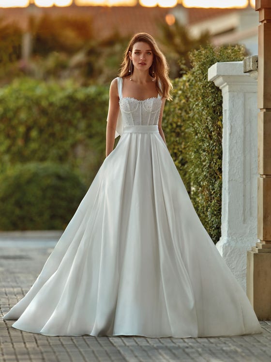 The classics reinvented, as in this princess-style satin dress with pockets on the skirt and a sensational trendy bodice with a visible structure and a play of pearls that perfectly match the bows on the straps framing a sensual square neckline. 