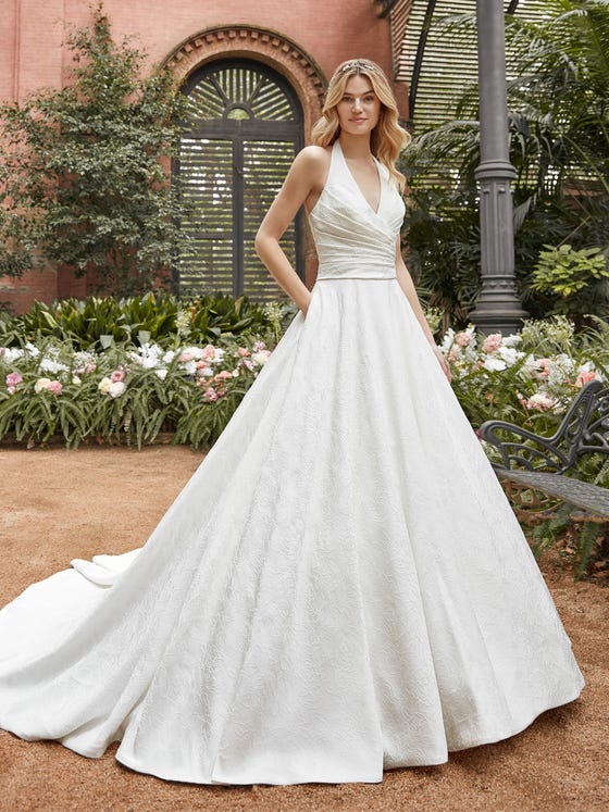 An elegant draped bodice and sensual halter neck lend an update to this classic gown crafted in textural brocade. With fashionable deep pockets and a circular cathedral train.  
