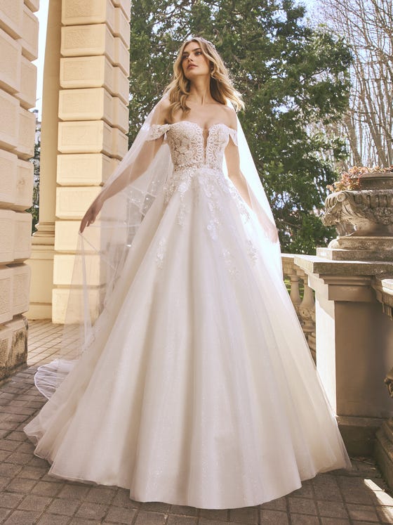 Look dazzling in this incredible princess dress in glimmery tulle with floral appliques and transparencies cascading down from the off-the-shoulder neckline with sleeves to the hip of the skirt. 