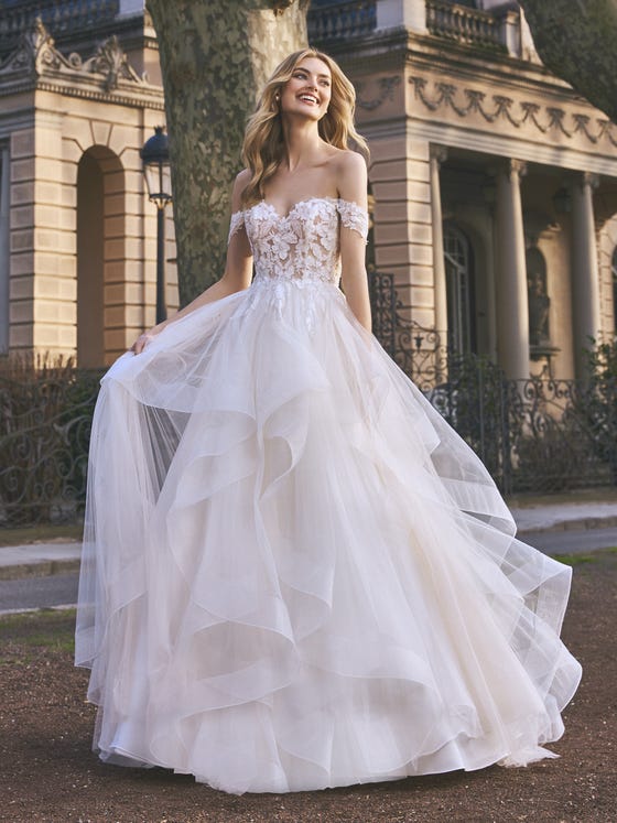 Ruffles and flowers. A design with loads of personality created by the asymmetrical volume of the crinkled tulle layers and a sensual semitransparent bodice with off-the-shoulder sleeves and a beautiful sweetheart neckline. 
