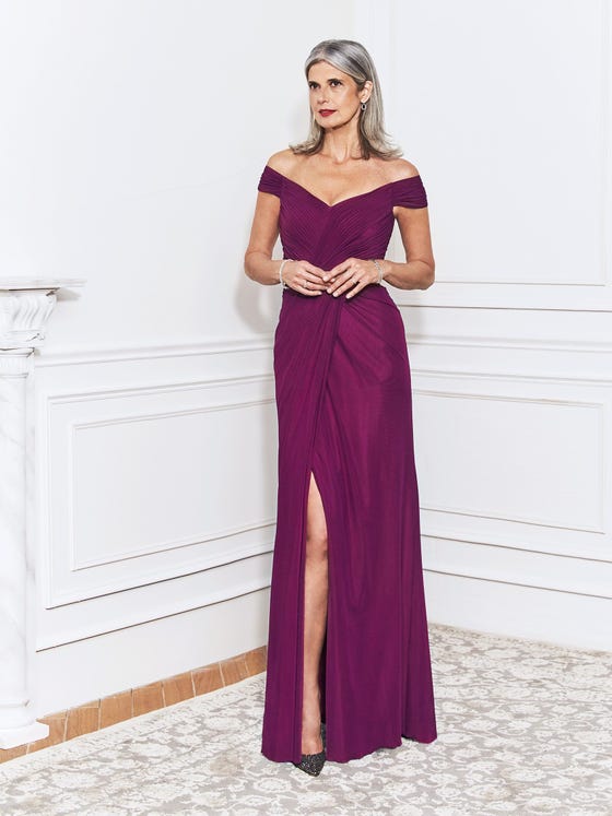 Elegant sheath gown in red violet stretch tulle, designed with fine drapery at the off-the-shoulder sleeves, twisted bodice, and layered sheath skirt that opens in a subtle slit. 